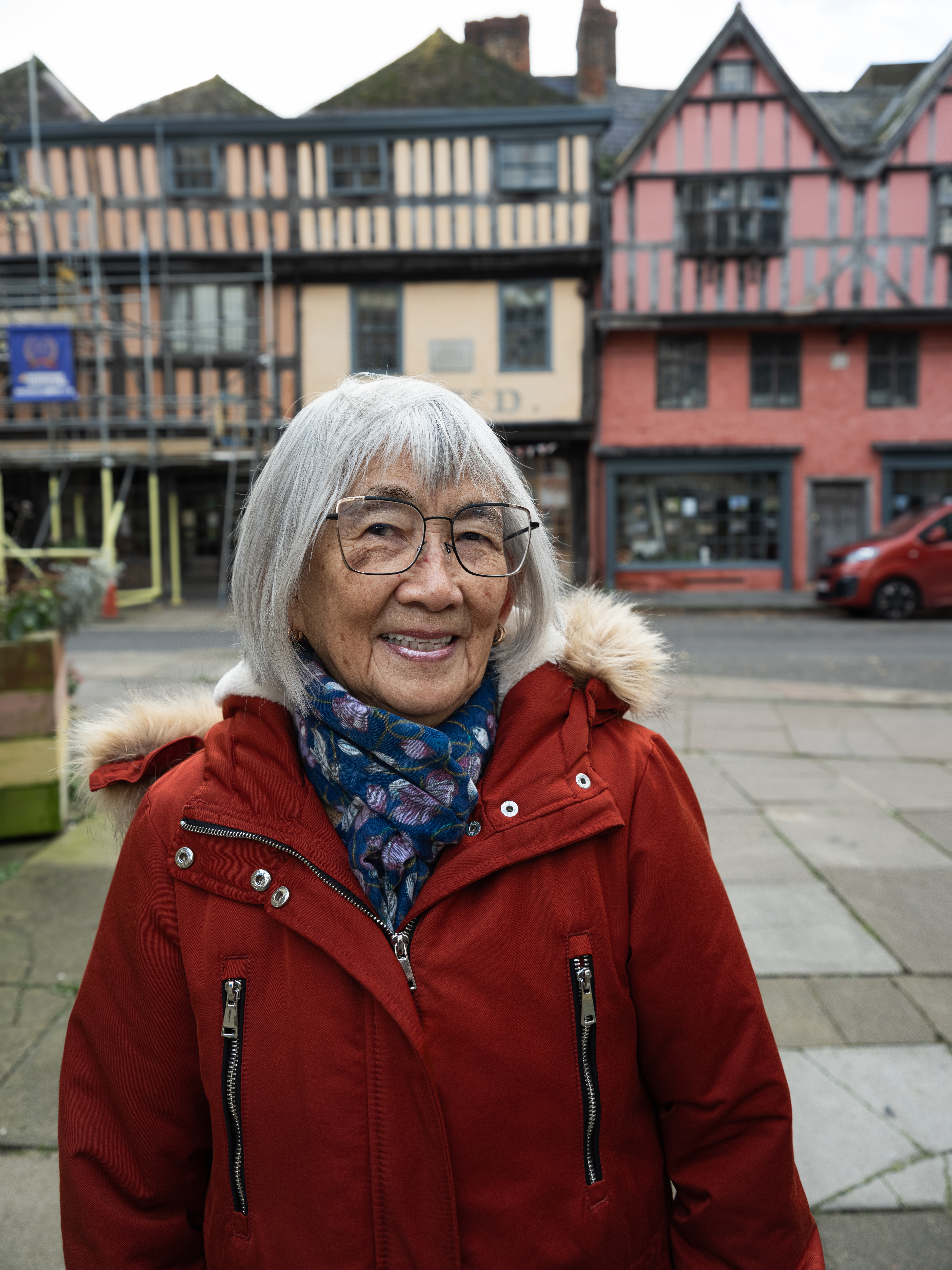 Alice she/her is an Asian woman from Sarawak in her 80s. She is photographed in Westgate street in front of the Gloucester Folk Museum where the family statue used to stand before it was vandalised and removed. 