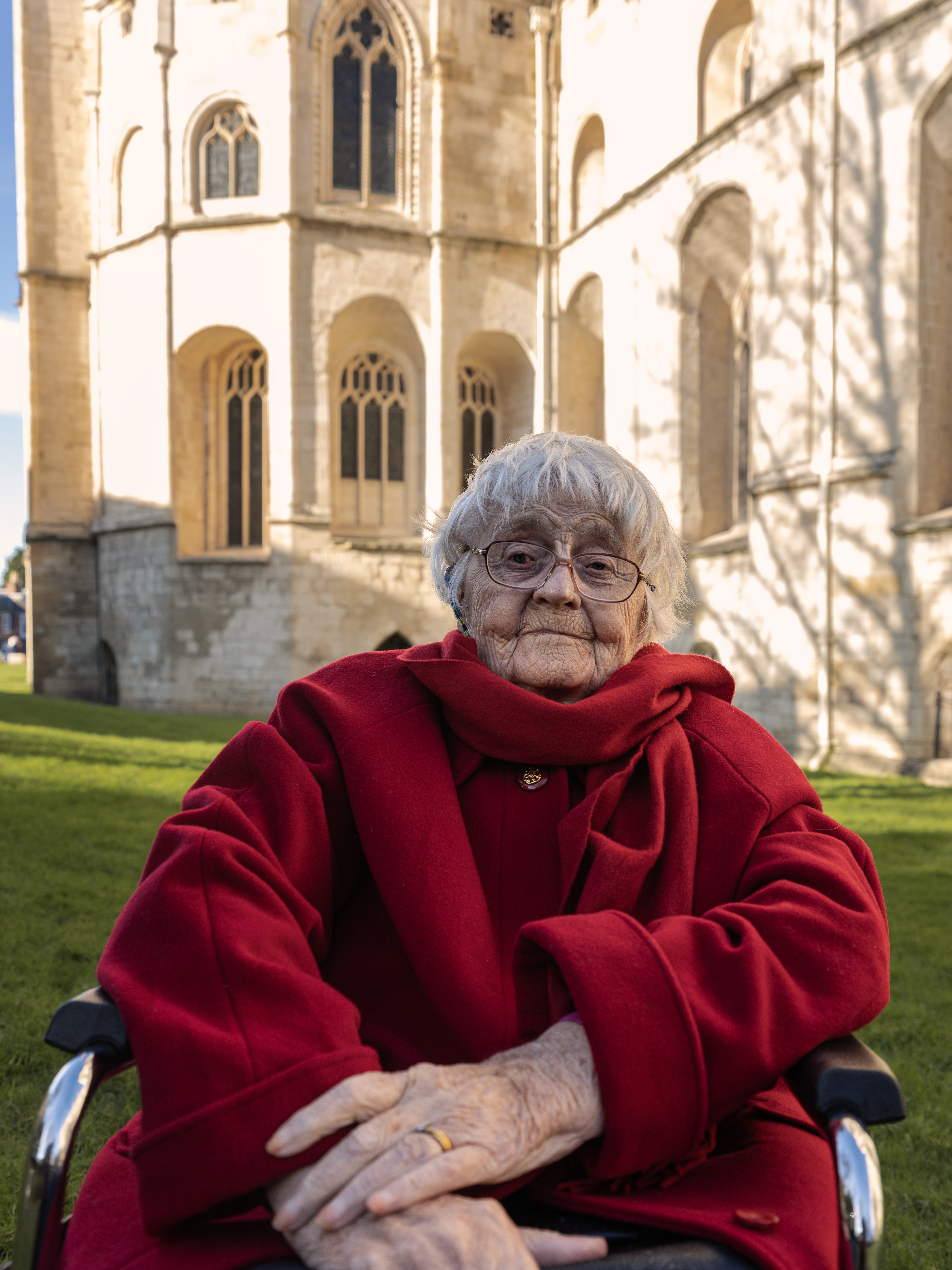 Jean she/her is a white woman in her mid 90s. She is photographed outside the Gloucester cathedral on a sunny winter's day afternoon. 