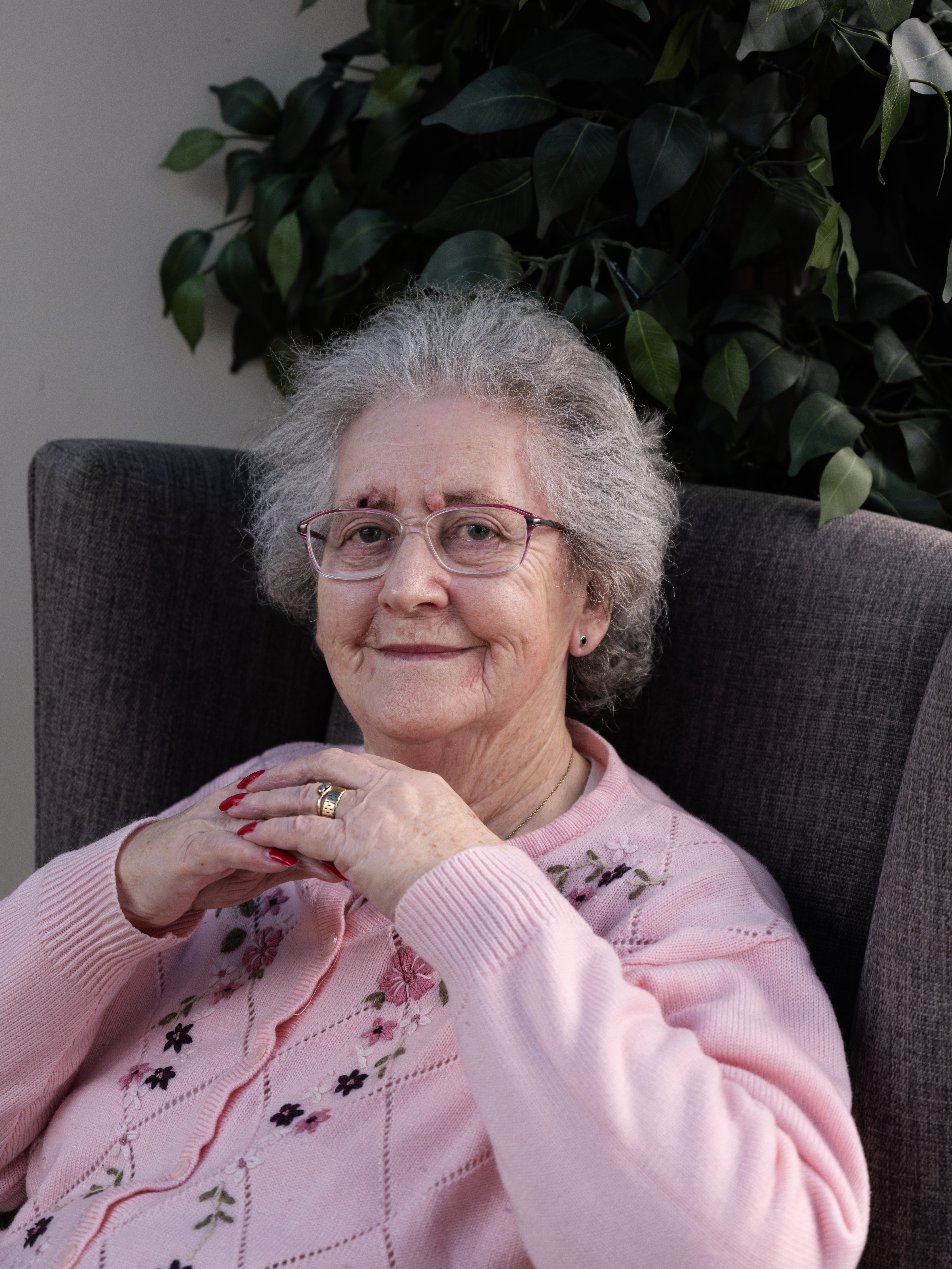 Shirley (she/her) is a white woman in her 70’s. She is pictured sat in dark slate grey armchair with a faux dark green plant behind her. At her residential care home in Gloucester. Shirley is seen sweetly smiling while posing for the camera. She loved having her picture taken. Her hands resting just below her chin. She has salt and pepper coloured hair which is styled off her face.  She is wearing a light baby pink button up knitted cardigan with dark pink and purple flowers embroidered down the front. She 