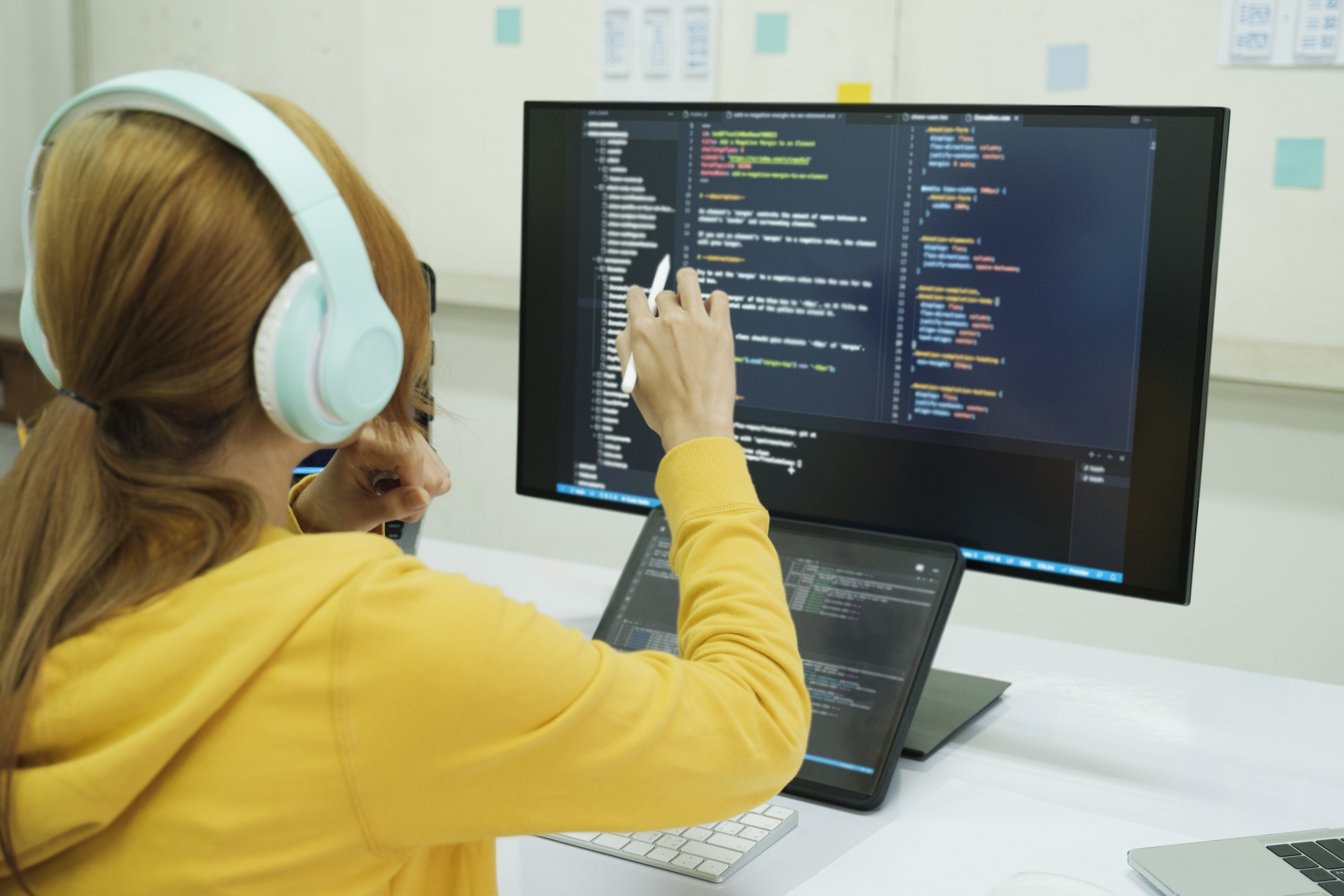 Photo of a young person in a yellow hooded jumper, wearing headphones, sat in front of a computer working on written programming code