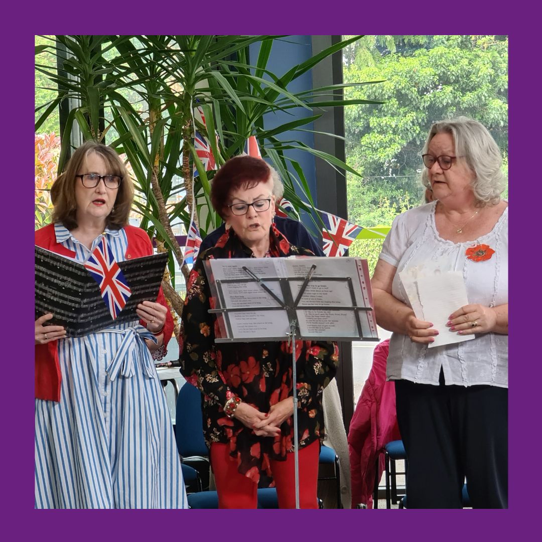A photo of three women singing in a choir wearing poppy pins with Union Flag bunting hanging in the background.