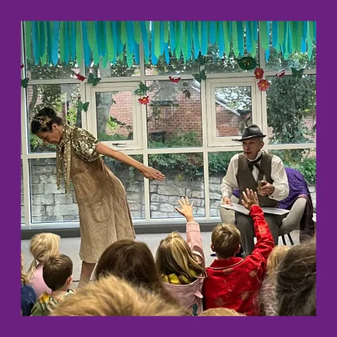 A storyteller sits in a chair in front of a group of children with raised hands while a dancer poses beside him