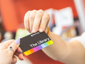 A smiling woman in a blue lanyard passes a library card into a receiving hand