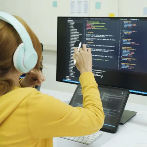 Photo of a young person in a yellow hooded jumper, wearing headphones, sat in front of a computer working on written programming code