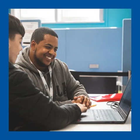 A photo of a man wearing a lanyard assisting a younger man with his laptop