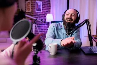 A person wearing headphones talking into a podcasting microphone while smiling.