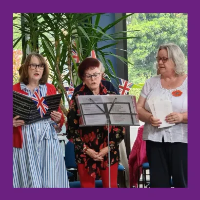 A photo of three women singing in a choir wearing poppy pins with Union Flag bunting hanging in the background.