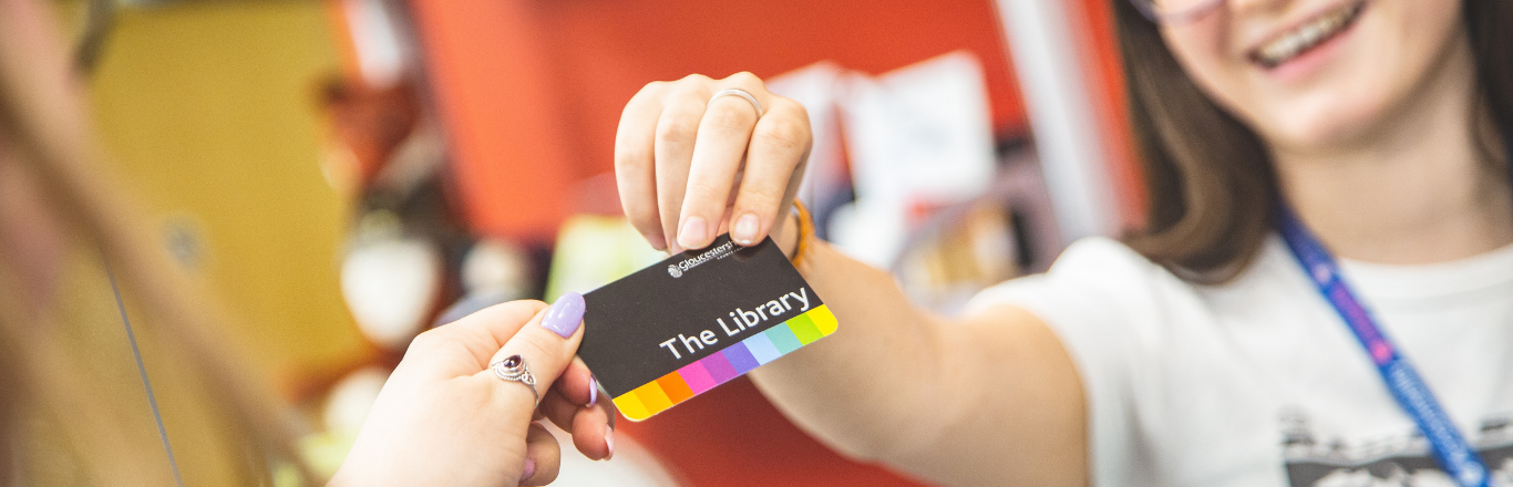 A smiling woman in a blue lanyard passes a library card into a receiving hand